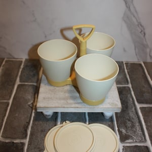 May include: A vintage cream-colored serving set with three bowls and a handle, resting on a small wooden stand. The set includes matching lids. The bowls and lids have a yellow accent color.