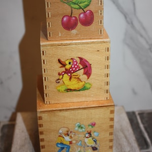 Może przedstawiać: Trzy ułożone drewniane pudełka z dekoracyjnymi ilustracjami. Górne pudełko przedstawia dwie czerwone wiśnie, środkowe pudełko ma kaczkę z różowym parasolem, a dolne pudełko przedstawia mężczyznę i ptaka. Pudełka są wykonane z jasnego drewna.