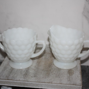 May include: Two white milk glass serving pieces. One is a sugar bowl with handles and a scalloped rim. The other is a creamer with a spout and handle. Both have a textured, hobnail pattern.