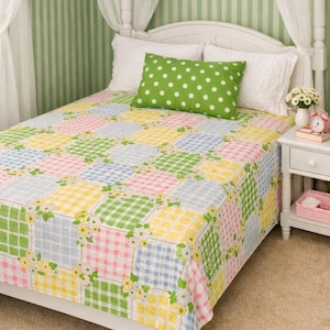 May include: A bedspread featuring a patchwork design of pastel pink, yellow, blue, and green gingham squares decorated with small white and yellow floral patterns. A single rectangular green pillow with white polka dots rests on the bed.