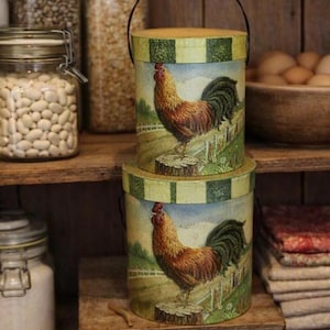 May include: Two cylindrical containers stacked vertically, each featuring a painted rooster standing on a tree stump in a field. The containers have a light yellow and green striped lid and a thin black metal handle attached to the top.