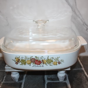 May include: White casserole dish with a clear glass lid. The dish features a colorful floral and fruit design around the rim, with the text "L'échalote" below the design. The dish is resting on a small, white stand.