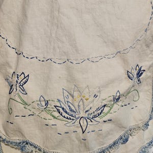 May include: White linen cloth with blue embroidered floral design. The embroidery features a large central flower with yellow accents, surrounded by smaller flowers and green stems. The edge is trimmed with blue crochet.