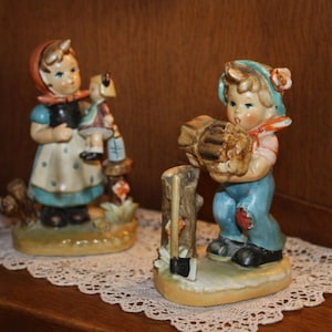 May include: Two hand-painted ceramic figurines. One depicts a child in a red headscarf holding a doll. The other shows a child in a blue cap carrying a bundle of logs next to a tree stump with an axe. Both figures stand on oval bases.