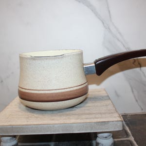 Op de afbeelding: Een vintage koffiepot in gespikkeld beige en bruin, met een donkerbruin handvat. De pot heeft een bruine streep in het midden en staat op een kleine, lichtgeleurde standaard. Het handvat is bevestigd met een zilverkleurig metalen stuk.