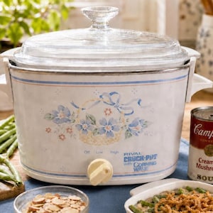 May include: A white Rival Crock-Pot with a clear glass lid. The slow cooker has a floral design with blue flowers and a basket. A can of Campbell's Cream of Mushroom Soup is next to the Crock-Pot. The control knob reads "Off, Low, High."