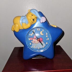 May include: A blue star-shaped analog alarm clock featuring a yellow Winnie the Pooh character wearing a blue nightcap on top. The clock face displays numbers one through twelve, a crescent moon, stars, and a graphic of Piglet sleeping on a cloud.