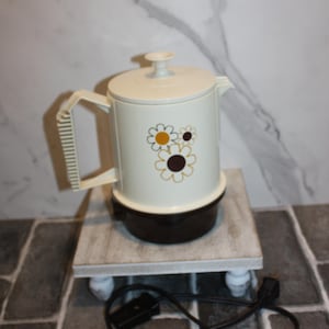 May include: A vintage cream-coloured electric coffee pot with a brown base and lid, decorated with daisy motifs in brown and yellow. The pot has a handle and spout. It sits on a small, white-legged wooden stand. A black power cord is visible.