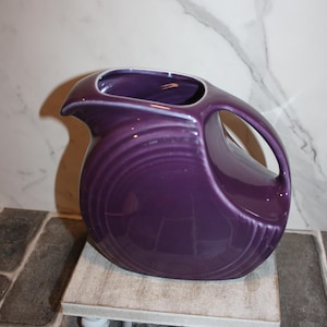 May include: A deep purple ceramic pitcher with a distinctive handle and spout. The pitcher has a rounded body with concentric ridges and sits on a small, white-legged stand. The background features a marble-patterned wall.