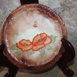 May include: A small, round porcelain plate with a decorative gold rim. The plate features a hand-painted design of two red poppies with green leaves and stems.
