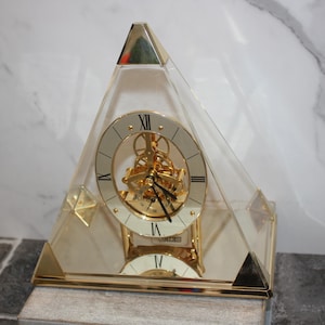 May include: A gold-toned pyramid clock with a clear acrylic casing. The clock face features Roman numerals and a visible mechanical movement. The clock is approximately 10 inches tall.