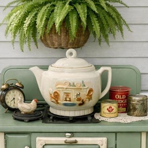 May include: A cream-colored ceramic teapot with a raised fireplace scene sits on a sage green stove. Beside it are a black alarm clock, a small white chicken figurine, a red Old Judge coffee tin, and a small yellow tin on a white lace doily.