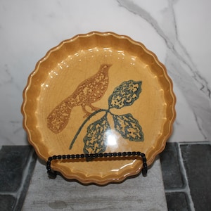 May include: A decorative, round, tan-colored plate with a scalloped edge. The plate features a brown bird and blue-green leaves design. The plate is displayed on a black metal stand.