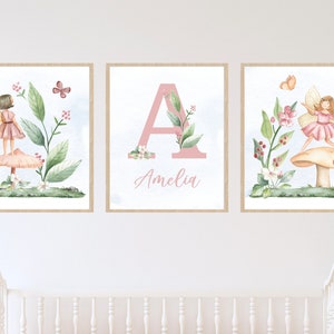 Peut inclure: Trois impressions aquarelle représentant une fée avec des ailes et une robe roses debout sur un champignon, une lettre A rose avec des feuilles vertes et le nom Amelia, et une fée avec des ailes et une robe roses debout sur un champignon avec des papillons qui volent autour.