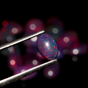 May include: A close-up of a dark purple and red oval opal gemstone held by tweezers. The opal is iridescent and has a speckled appearance.
