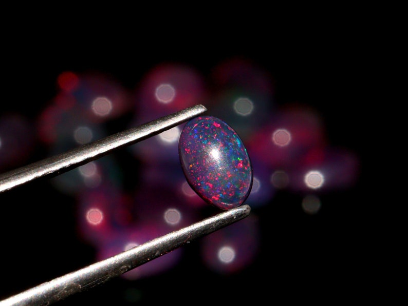 May include: A close-up of a dark purple and red oval opal gemstone held by tweezers. The opal is iridescent and has a speckled appearance.