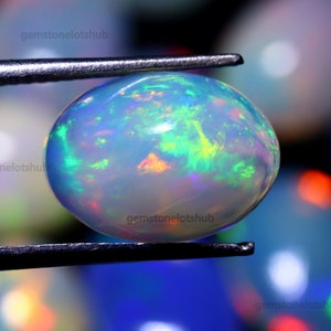 May include: A white opal gemstone with a rainbow of colors, including green, blue, and red, is held in a pair of tweezers. The opal is oval shaped and has a smooth, polished surface.