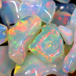 May include: A close-up of a pile of iridescent white opal stones. The stones are smooth and have a variety of shapes and sizes. The opals are reflecting light, creating a rainbow of colors.