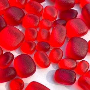May include: A collection of smooth, red sea glass pieces. The pieces vary in size and shape, with rounded edges and a frosted appearance. The color ranges from bright red to deep burgundy, creating a vibrant display.