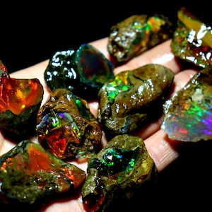 May include: A collection of rough opal stones with iridescent colours, including green, blue, red, and orange. The stones are in various shapes and sizes and are set in a dark brown matrix.