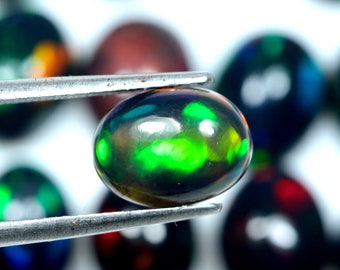 May include: A black opal gemstone held by tweezers, displaying vibrant green and yellow flashes. The polished oval stone is the focal point, with other opals slightly out of focus in the background. The gemstone is a beautiful example of natural opal.