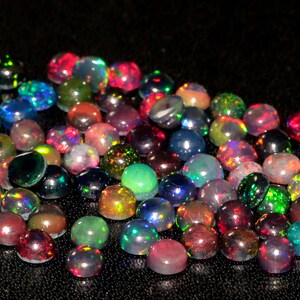May include: A collection of round, iridescent opal gemstones in various shades of green, blue, red, and black. The opals are arranged in a loose pile on a black surface.