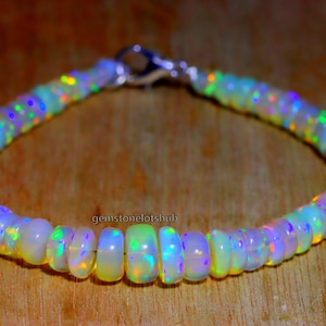 Puede incluir: Un brazalete de cuentas de ópalo blanco con un cierre plateado. Las cuentas son iridiscentes y muestran destellos de color, incluyendo verde, azul y morado.