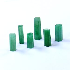 Puede incluir: Siete cristales de piedras preciosas verdes de diferentes alturas están dispuestos sobre una superficie blanca. Los cristales tienen forma cilíndrica con tapas facetadas y exhiben un color verde vibrante y translúcido. Las piedras preciosas probablemente se utilizan para joyería o fines decorativos.