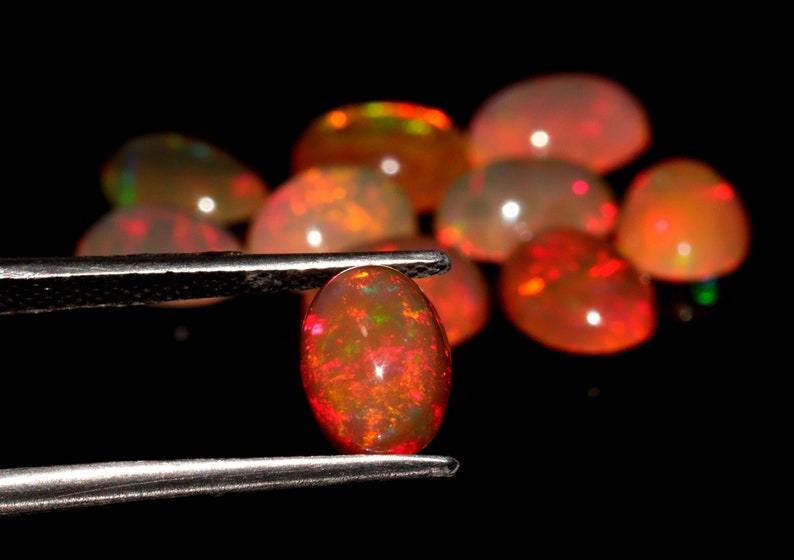 May include: A close-up of a red opal gemstone held in tweezers. The opal is oval-shaped and has a fiery red color with flashes of green and yellow.