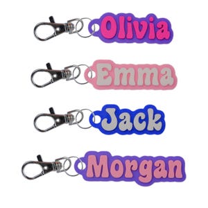 May include: Four colorful personalized keychains with metal rings and clips. The names Olivia, Emma, Jack, and Morgan are printed in white letters on a pink, purple, blue, and purple background.