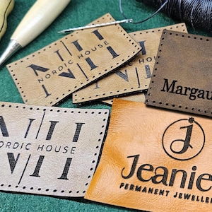 May include: Four leather labels with different designs. The first two labels are brown and feature the text "NORDIC HOUSE" and Roman numerals. The third label is brown and features the text "Margaux H.D.". The fourth label is light brown and features the text "Jeanie's PERMANENT JEWELLERY" and a stylised letter "J".