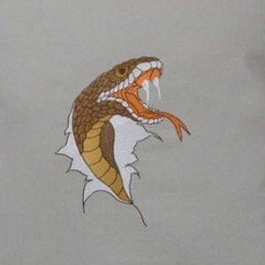 Puede incluir: Una almohada beige con un diseño de serpiente bordada en marrón y naranja. La serpiente está representada con la boca abierta y los colmillos visibles, como si fuera a atacar. La serpiente está bordada sobre un fondo blanco, que está rodeado de una tela beige.