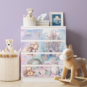 May include: A white dresser with four drawers, each adorned with a watercolor illustration of mermaids, sea creatures, and a castle. The top of the dresser displays toys and a framed picture. A rocking horse and a basket of toys are in the foreground.