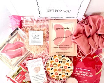 Puede incluir: Una caja de regalo rosa llena de artículos de belleza y cuidado personal. Incluye mascarillas para los ojos, una goma para el pelo, té, una vela, un posavasos con diseños de café y cruasanes, y una mascarilla peel-off de sandía. La caja tiene el texto "JUST FOR YOU".