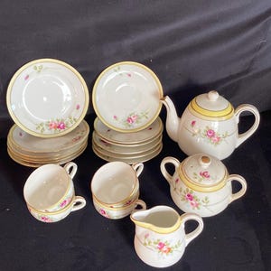 May include: A vintage tea set featuring a teapot, sugar bowl, creamer, cups, and saucers. The white porcelain set is decorated with pink roses, yellow trim, and gold accents. The set includes multiple cups and saucers.