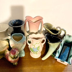 May include: A collection of vintage ceramic vases in various colors and styles, including a pink heart-shaped vase, a blue and white floral vase, a green pitcher, and a black and white vase.