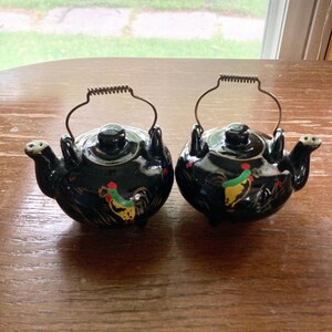 May include: A set of two black ceramic salt and pepper shakers shaped like teapots with rooster designs. The shakers have metal wire handles and are decorated with black, red, green, and yellow paint.