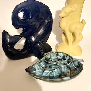 May include: A collection of ceramic decorative items. Includes a glossy black whale sculpture, a light yellow vase with a deer design, and a leaf-shaped dish with blue and green marbling.