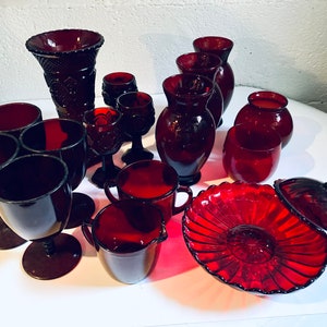 May include: A collection of ruby red glass items, including vases, bowls, cups, and a lidded dish. The items are arranged on a white surface.