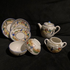 May include: A miniature porcelain tea set with a whimsical design. The set includes a teapot, creamer, sugar bowl, saucers, and cups. Each piece features a colorful illustration of people and scenes, with blue and gold accents. The set is likely vintage.