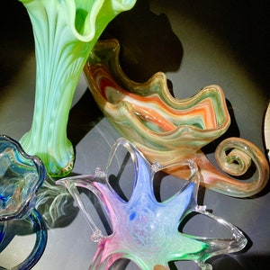 May include: A collection of colorful glass art pieces. Includes a tall, ruffled vase in shades of green and blue, a star-shaped bowl with pink, blue, and green hues, and a swirled bowl with orange and white tones.