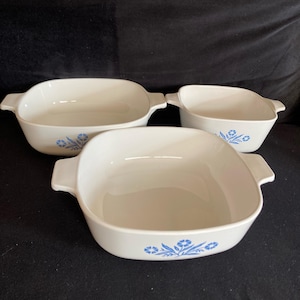 May include: Three white, square-shaped casserole dishes with handles. Each dish features a blue floral design on the side. The dishes are of varying sizes and are arranged on a dark surface.