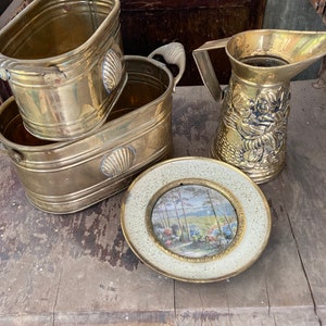 May include: Two brass planters with shell-shaped handles, a brass pitcher with an embossed floral design, and a small brass picture frame with a landscape scene.