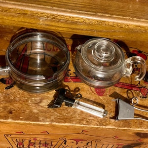 May include: A vintage glass teapot and a double-walled glass pot with a clear plastic handle. The teapot has a silver lid and a silver band around the bottom. The pot has a silver band around the top and bottom. There are also two silver metal tongs and a silver metal handle.