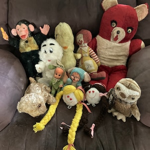 May include: A collection of vintage plush toys, including a monkey, a bear, a sloth, a clown, and several dolls. The toys are arranged on a brown leather couch.