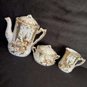 May include: A white porcelain tea set with gold accents. The set includes a teapot, a sugar bowl with a lid, and a creamer. Floral and leaf patterns decorate the pieces. The set is on a black surface.