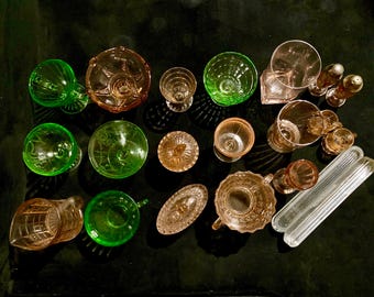 May include: A collection of vintage glass dishes in green and pink hues. The dishes are arranged on a black surface and include bowls, cups, and a pitcher.