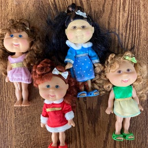 May include: Four Cabbage Patch Kids dolls in different outfits. The dolls are wearing a pink dress, a red dress, a blue dress, and a green dress. They all have curly hair and big smiles.