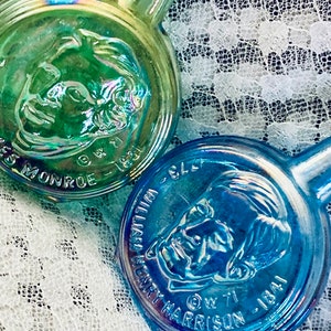 May include: Two iridescent glass bottle stoppers featuring the portraits of James Monroe and William Henry Harrison. The stoppers are blue and green and have the text "James Monroe - 1821" and "William Henry Harrison - 1841".