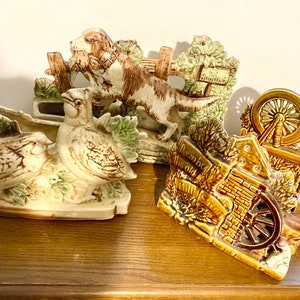 May include: Four ceramic planter figurines with a country cottage theme. The planters feature a dog chasing a bird, two birds, a cottage with a wheel, and a spinning wheel. The planters are painted in shades of brown, green, and white.